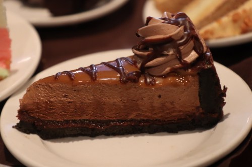 A slice of chocolate cheesecake topped with whipped cream and drizzled with caramel sauce on a white plate.