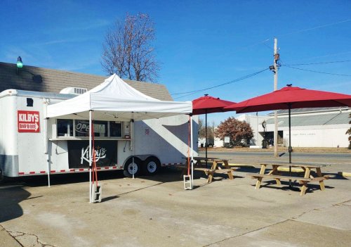 A food truck with a sign reading "Kilby's Bar-B-Que," featuring outdoor seating under red umbrellas.