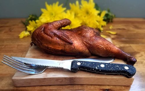 A roasted chicken on a wooden cutting board, accompanied by a knife and fork, with yellow flowers in the background.