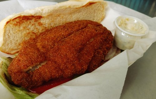 A crispy fried fish fillet on a bun with lettuce, ketchup, and a side of tartar sauce in a takeout container.