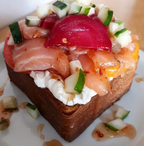 A square piece of bread topped with cream cheese, salmon, tomatoes, cucumbers, and a drizzle of sauce.