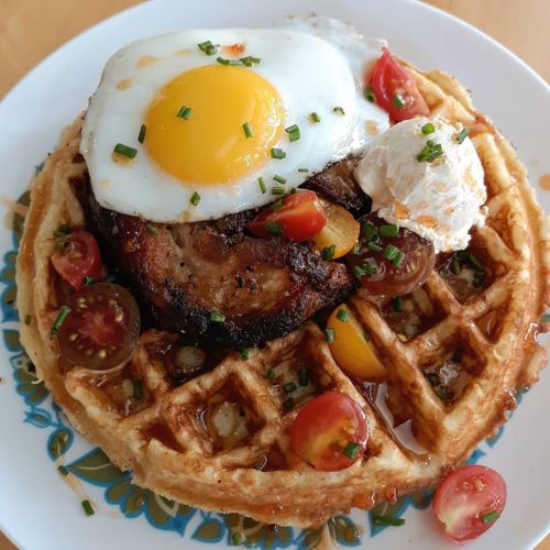 A golden waffle topped with a fried egg, poached egg, sliced tomatoes, and a piece of meat, drizzled with syrup.