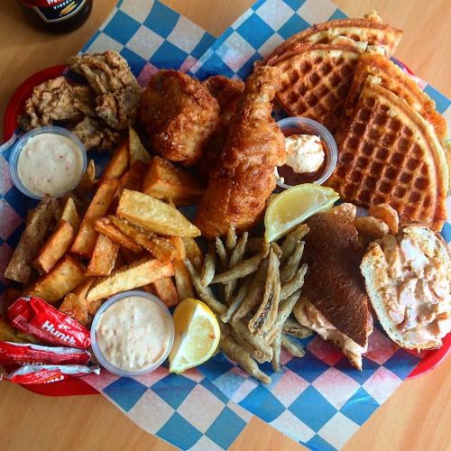 A vibrant platter featuring fried chicken, waffles, seasoned fries, fish, and dipping sauces on a checkered paper.