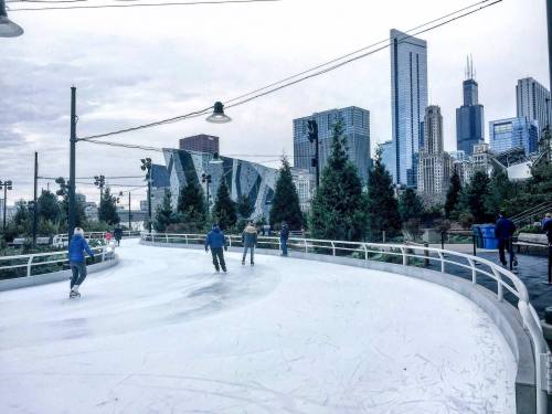 People ice skating on a rink with a city skyline in the background, featuring tall buildings and trees.