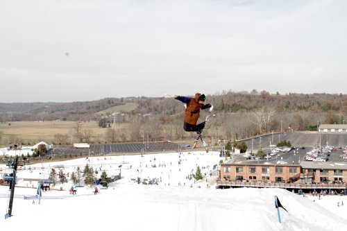 A skier performs a jump over a snowy slope with a resort and parking lot in the background.