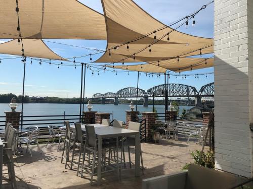 Outdoor dining area with string lights, overlooking a river and a bridge under a blue sky.