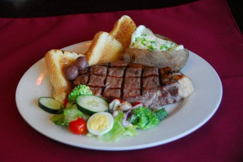 A plate featuring grilled steak, toast, a baked potato, and a fresh vegetable salad with sliced eggs.