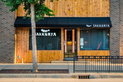 Exterior of Sakanaya restaurant featuring wooden paneling, a black awning, and large windows.