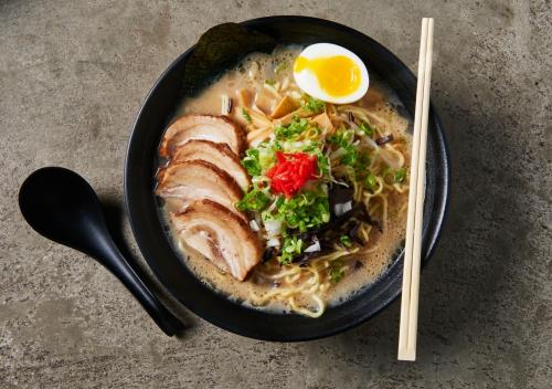 A bowl of ramen with sliced pork, green onions, bamboo shoots, a soft-boiled egg, and a slice of nori on a textured surface.