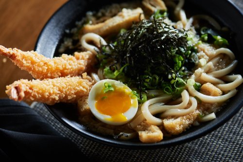 A bowl of udon noodles topped with green onions, seaweed, a soft-boiled egg, and two crispy shrimp tempura.