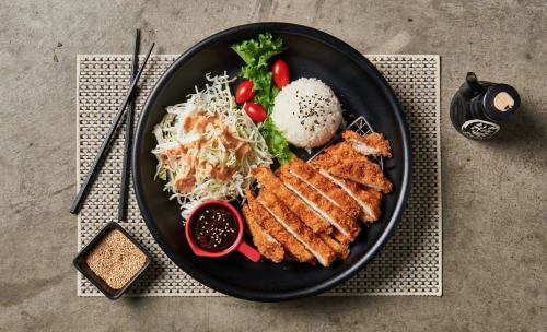 A black plate with crispy chicken, rice, salad, cherry tomatoes, and dipping sauce, served on a textured mat.