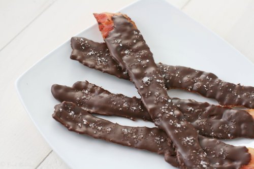 Chocolate-covered bacon strips sprinkled with sea salt on a white plate.