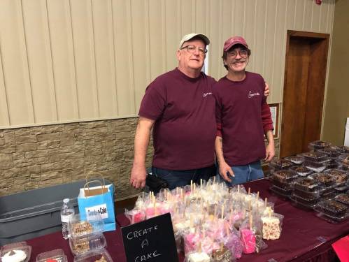 Two men in maroon shirts stand behind a table filled with desserts and a sign that says "Create a Cake."