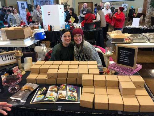 Two smiling women sit behind a table filled with neatly stacked boxes at a bustling event with people in the background.