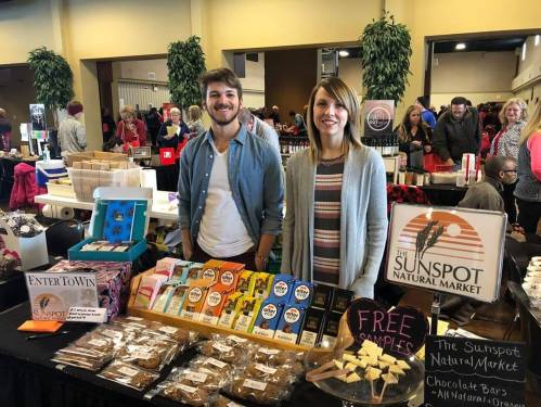 Two smiling vendors stand behind a table filled with snacks and products at a market, promoting The Sunspot Natural Market.