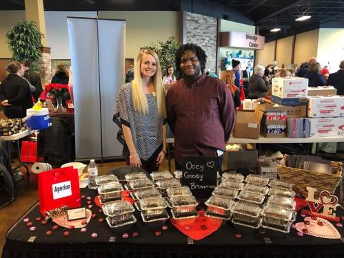 Two vendors stand behind a table filled with brownies, smiling at a community event. Boxes and people are in the background.