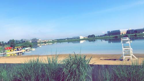 A calm lake with boats and a lifeguard chair, surrounded by green grass and buildings in the background under a clear sky.