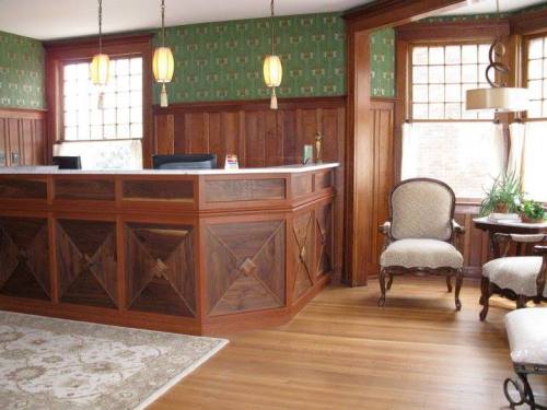 A wooden reception desk in a warmly decorated room with green wallpaper, vintage chairs, and natural light.