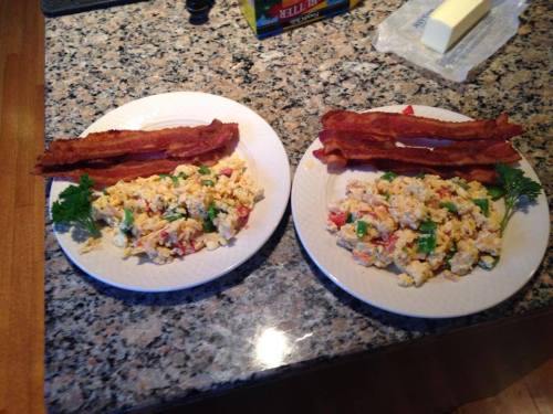 Two plates of scrambled eggs with vegetables and strips of bacon, garnished with parsley, on a granite countertop.