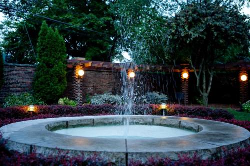 A circular stone fountain with water spraying up, surrounded by lush greenery and softly lit by garden lights.