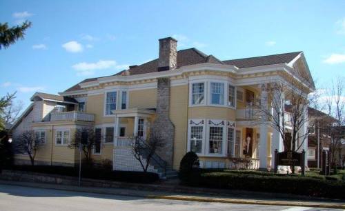 A large, elegant yellow house with a stone chimney, surrounded by trees and a well-maintained lawn under a blue sky.