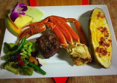 A gourmet plate featuring crab legs, a beef patty, baked potato, mixed vegetables, and a slice of melon with a flower.