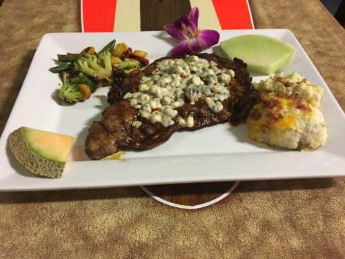 A plated meal featuring a grilled steak topped with cheese, accompanied by vegetables, mashed potatoes, and melon.