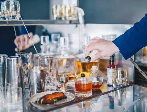 A hand pours a cocktail into a glass on a bar, surrounded by various drinkware and a plate of food.