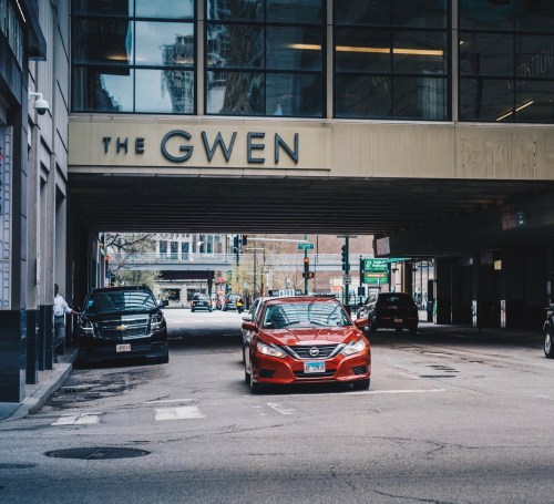A red taxi drives under a building sign reading "The Gwen" in a city street scene.