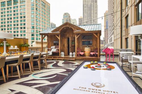 A rooftop terrace at The Gwen Hotel features a wooden cabin, seating, and a curling rink with city skyline views.