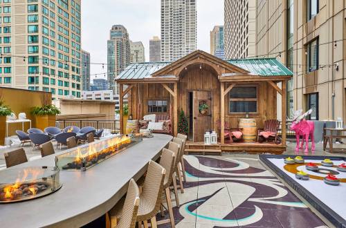 A rooftop space featuring a wooden cabin, modern seating, a fire pit, and city skyline views.