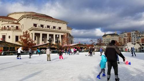 A bustling outdoor ice skating rink with people skating, surrounded by buildings and autumn trees under a cloudy sky.