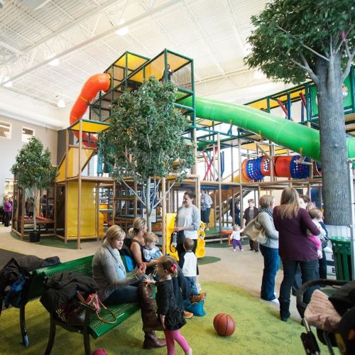 Indoor playground with colorful slides, climbing structures, and families enjoying the space. Trees and benches are visible.