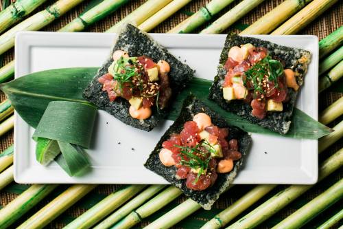 Three sushi bites on a white plate, topped with tuna, avocado, and sauces, surrounded by bamboo leaves.