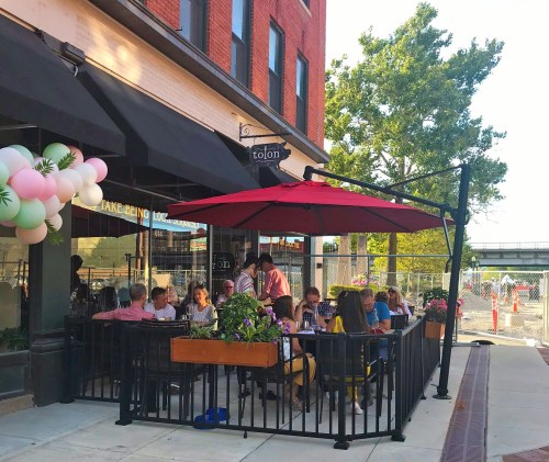 Outdoor dining area with people at tables under umbrellas, surrounded by greenery and balloons, near a brick building.
