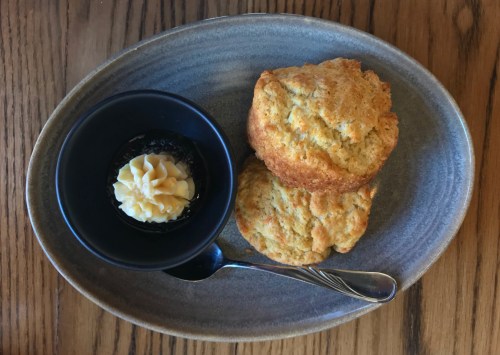A plate with two golden biscuits and a small bowl of creamy butter on the side, accompanied by a spoon.
