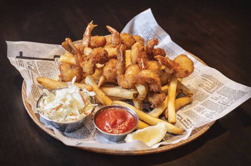 A plate of golden fried shrimp served with fries, coleslaw, cocktail sauce, and lemon wedges on a newspaper-lined tray.