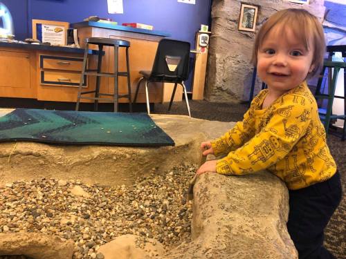 A smiling toddler in a yellow shirt plays at a sand and pebble area in a colorful indoor space.