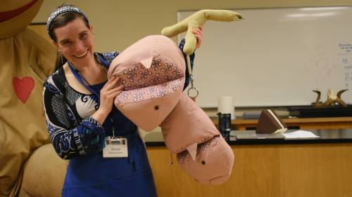 A smiling woman in a blue patterned shirt holds a large, plush anatomical model of a penis in a classroom setting.