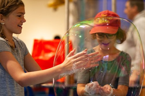 Two children play with a large soap bubble, smiling and reaching out to touch it in a colorful indoor setting.