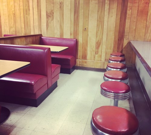 A retro diner interior featuring red booths, wooden walls, and chrome stools along a counter.