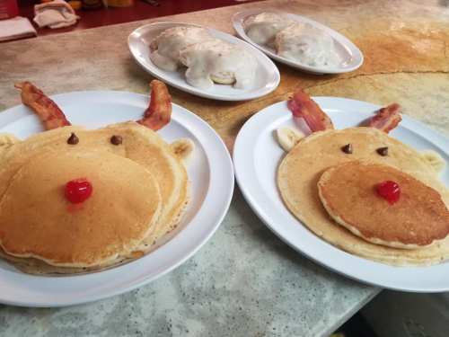 Two plates of pancakes decorated as reindeer faces, with bacon antlers and cherry noses, alongside plates of biscuits and gravy.