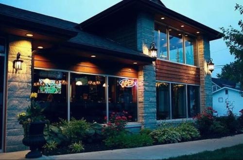 A cozy restaurant exterior at dusk, featuring large windows, an "Open" sign, and lush landscaping.