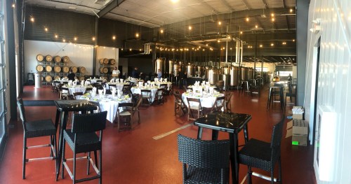 A spacious restaurant with tables set for dining, string lights overhead, and brewing equipment in the background.