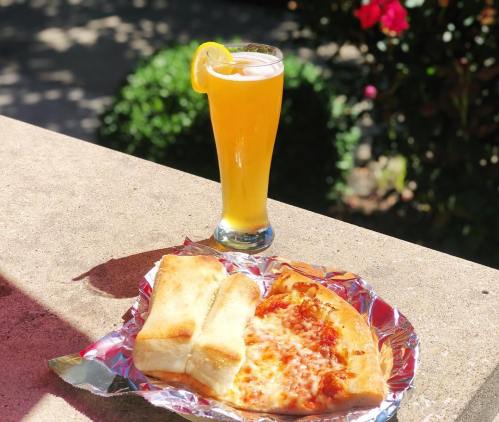 A glass of yellow beverage with a slice of cheesy pizza and breadsticks on a foil-lined plate, set outdoors.