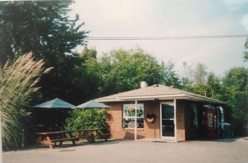 A small brick building with outdoor seating, surrounded by greenery, featuring a Coca-Cola vending machine.
