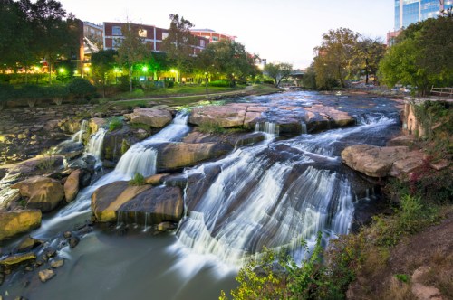 A serene waterfall cascades over rocks, surrounded by greenery and city lights at dusk.