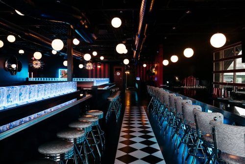 A stylish bar interior with a black and white checkered floor, illuminated by round pendant lights and a glowing blue bar.