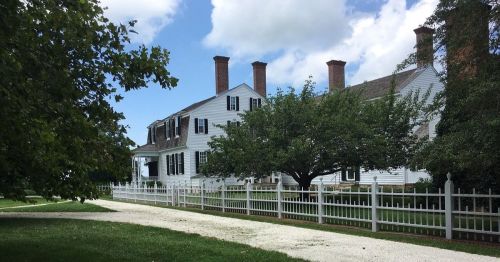 A historic white house with multiple chimneys, surrounded by green trees and a white picket fence under a blue sky.