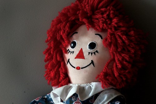 A close-up of a vintage rag doll with bright red curly hair, a cheerful smile, and a patterned dress.